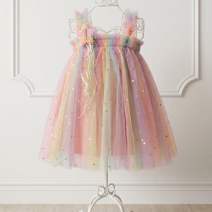 Puede incluir: Un vestido sin mangas hecho de capas de tul arcoíris en tonos pastel con detalles de estrellas doradas en toda la tela. El corpiño tiene un escote fruncido y una aplicación decorativa en forma de estrella rosa con el número uno y cintas colgantes.
