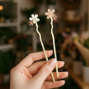 May include: Two gold-colored hair sticks, each adorned with a delicate floral design. One features white and burgundy flowers, the other pink and burgundy. The floral accents add a touch of elegance to these hair accessories.