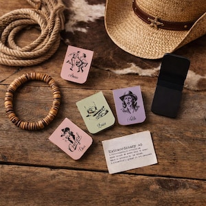 May include: A collection of colorful, rectangular bookmarks with illustrations and names, including "Amer," "Louis," and "Felle." A cowboy hat, a wooden bracelet, and a piece of paper with the word "Extraordinary" are also present on a wooden surface.