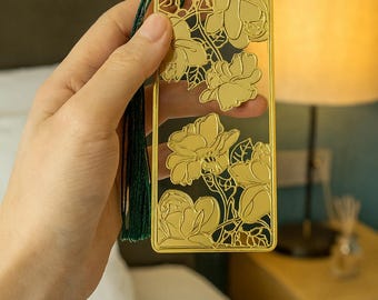 Könnte beinhalten: Goldfarbener Metall-Lesezeichen mit floralem Design. Das Lesezeichen hat eine rechteckige Form mit ausgeschnittenen Blumen und Blättern. Eine grüne Quaste hängt oben. Das Lesezeichen wird in einer Hand gehalten.
