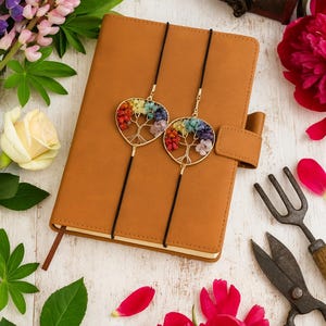 May include: A brown leather journal with a pen loop and two tree of life pendants with colorful gemstones. The pendants hang on black cords. The image is surrounded by flowers and gardening tools.