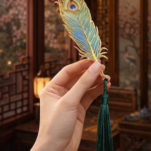 Può includere: Segnalibro decorativo a forma di piuma di pavone. Il contorno è in metallo dorato, riempito di smalto blu e turchese. Un nappa verde scuro pende dal basso. Il segnalibro è tenuto in mano.