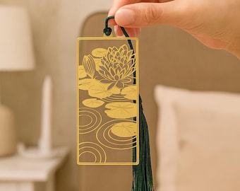 Könnte beinhalten: Goldfarbener Metall-Lesezeichen mit einem ausgeschnittenen Design einer Seerose und Seerosenblättern. Das Lesezeichen hat eine rechteckige Form mit einer grünen Quaste. Das Design umfasst eine detaillierte Blume und Wassermuster.