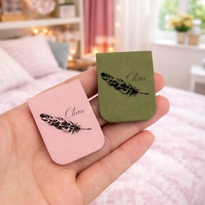 May include: Two small rectangular bookmarks, one pink and one olive green, are held in a hand. Each bookmark features a black feather design and the name "Clara" in script. The background includes a bed and a shelf.