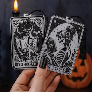 Puede incluir: Dos marcapáginas en blanco y negro estilo tarot con figuras de esqueletos leyendo libros. Un marcapáginas dice "THE READER" y el otro "THE READER". Cada uno tiene una borla negra y un borde decorativo. Tema de Halloween.
