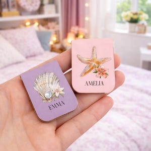 Puede incluir: Dos marcadores magnéticos personalizados. Uno es lavanda con una concha y el nombre "EMMA". El otro es rosa con una estrella de mar y el nombre "AMELIA". Ambos tienen detalles florales.