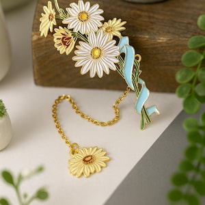 Puede incluir: Un broche de margarita dorado con pétalos blancos y centro amarillo, acentuado por una cinta azul claro. Un dije de margarita a juego cuelga de una cadena. Un accesorio floral.