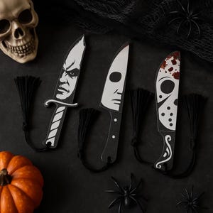 May include: Three Halloween-themed knife-shaped bookmarks with black tassels. Each bookmark features a different horror movie character's face in black and white. The background is black with a skull, spiders, and a pumpkin.