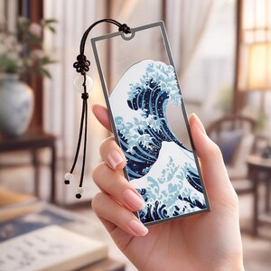 May include: A rectangular bookmark with a wave design in blue and white, with a black tassel and decorative beads. The bookmark is held in a hand, showcasing the intricate artwork and elegant design.