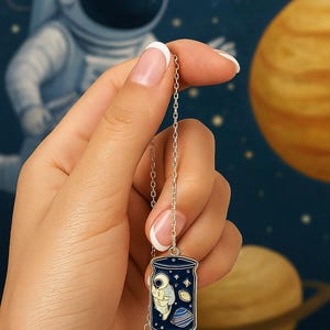 Könnte beinhalten: Silberfarbene Halskette mit einem Anhänger, der einen Astronauten in einem Glas, Planeten und Sterne zeigt. Der Anhänger ist blau, weiß und türkis. Der Hintergrund zeigt ein verschwommenes Bild eines Astronauten und Planeten.