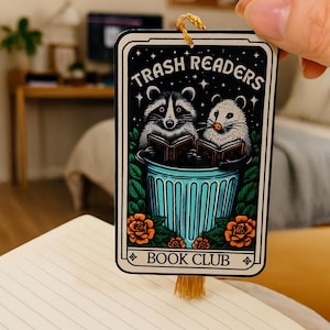 May include: A rectangular bookmark with the text "Trash Readers Book Club" features illustrations of two raccoons reading books in a blue trash can. The bookmark has a gold tassel and is held over an open notebook.