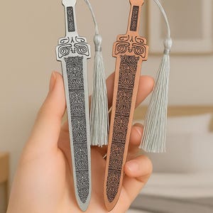 Può includere: Due segnalibri decorativi in metallo a forma di spada. Uno è color argento, l'altro color rame. Entrambi hanno intricati motivi incisi e un nappa. I segnalibri sono tenuti in una mano.