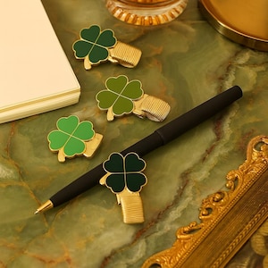 Op de afbeelding: Vierbladige klaver papieren clips in verschillende tinten groen en goud. Een zwarte pen ligt tussen de clips op een groen marmeren oppervlak. De klaverclips zijn gerangschikt in de buurt van een stapel crèmekleurig papier.