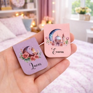 Pode incluir: Dois marcadores retangulares com desenhos islâmicos. Um é lavanda com "Lucas" e o outro rosa com "Emma". Ambos apresentam uma lua crescente, uma mesquita e motivos florais. O fundo é um quarto desfocado.