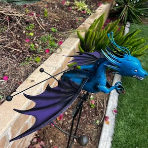 May include: A blue metal dragon garden stake with purple wings. The dragon has a long neck and tail, and is perched on a black metal stand. The wings are a deep purple with a gradient effect. The dragon is in a garden setting.