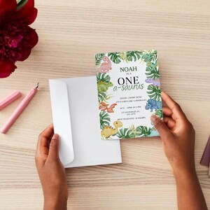 May include: A colorful invitation with the text "Noah is a One a-saurus" surrounded by dinosaur illustrations and tropical leaves. The invitation is held with a white envelope, a pink pen, and a red flower on a wooden surface.
