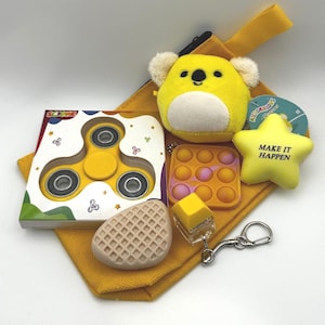Pode incluir: Um conjunto de brinquedos e acessórios amarelos, incluindo um fidget spinner, um peluche de coala, uma bola de stress em forma de estrela com o texto "MAKE IT HAPPEN", um brinquedo pop-it, um sabonete em forma de waffle e uma bolsa amarela. Em fundo branco.
