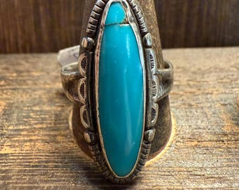 May include: A silver ring with an oval turquoise stone. The stone is set in a detailed, ornate silver band. The ring is displayed on a wooden ring holder, showcasing the jewelry's design and craftsmanship. The ring is a piece of jewelry.