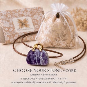 May include: A purple amethyst stone carved into a purse shape with a gold-tone metal bail, suspended on an 18-inch brown leather cord. The necklace is displayed with a floral-patterned gift bag and a card labeled Designs4Seasons.