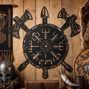 Peut inclure: Une décoration murale en métal noir représentant un symbole Vegvisir circulaire central, flanqué de deux haches croisées et surmonté d'une flèche. Le design présente des détails découpés complexes et des motifs géométriques sombres.