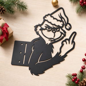 May include: A black metal wall art piece featuring a cutout silhouette of a character wearing a Santa hat, with a grumpy facial expression and one hand raised with a middle finger extended. The design includes a rectangular base with three slots.