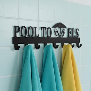 May include: A black metal wall-mounted rack with seven hooks, featuring the text "POOL TOWELS" and a graphic of an umbrella and flip-flops. Two teal towels and one yellow towel are hanging from the hooks on this decorative storage piece.