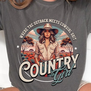 Peut inclure: T-shirt gris foncé avec un graphisme représentant une femme portant un chapeau de cow-boy, le texte "COUNTRY Girl" et la phrase "WHERE THE OUTBACK MEETS COWGIRL GRIT". Le t-shirt est de style décontracté.