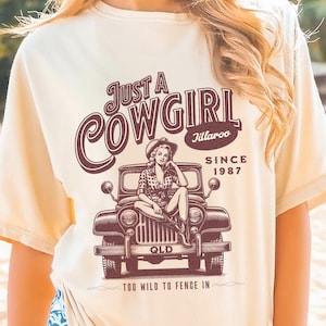 Peut inclure: T-shirt crème avec un motif marron représentant une cowgirl dans une jeep. Le texte indique "Just a Cowgirl Since 1987" et "Too Wild to Fence In". Le t-shirt est porté sur une plage.