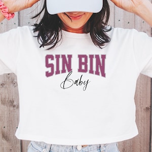 May include: White crop top with the text "SIN BIN Baby" in a purple collegiate font. The model is wearing a white baseball cap and light blue jeans. A pink scrunchie is on the model's wrist.