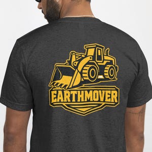 May include: A dark charcoal gray short-sleeve t-shirt featuring a yellow graphic on the back. The graphic depicts a construction vehicle with the word EARTHMOVER printed in bold capital letters inside a shield-shaped border beneath the machine.