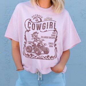 Peut inclure: T-shirt rose clair avec un motif marron d'une cowgirl sur un quad, avec le texte "Outback Cowgirl" et "Since 1984". Le t-shirt a des manches courtes et un col rond.