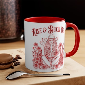 Peut inclure: Mug en céramique blanche avec intérieur et anse rouges. L'illustration rouge représente une femme en chapeau de cow-boy, entourée de fleurs, avec le texte "Rise & Buck Off". Une cuillère et un macaron sont au premier plan.