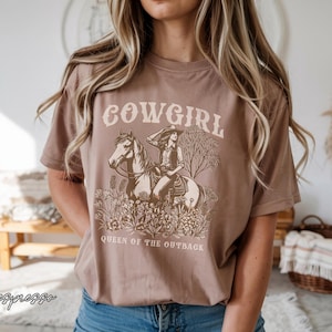 Peut inclure: T-shirt marron avec un motif de cowgirl à cheval, entourée de fleurs. Le mot "COWGIRL" est au-dessus de l'image, et "QUEEN OF THE OUTBACK" en dessous. Le mot "espresso" est écrit en cursive en bas à gauche du t-shirt.