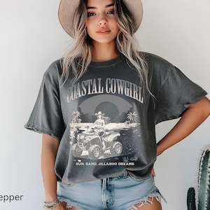 Peut inclure: T-shirt gris foncé avec l'inscription "COASTAL COWGIRL" au-dessus d'une illustration d'une cowgirl sur un quad. Le texte "SUN, SAND, JILLAROO DREAMS" est en dessous. Le t-shirt est porté par une personne.