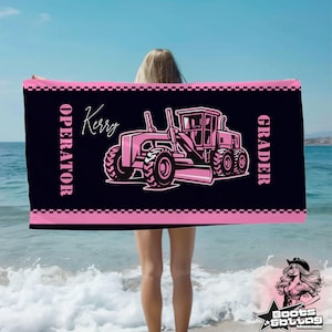 May include: A black rectangular beach towel featuring a pink graphic of a motor grader construction vehicle. The text "OPERATOR" and "GRADER" are printed in pink on the sides, with the name "Kerry" in a script font above the vehicle.