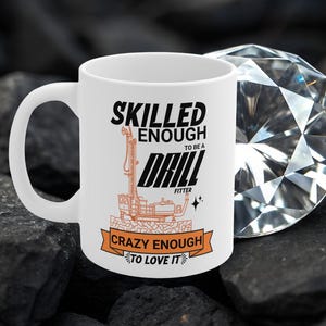 Op de afbeelding: Witte keramische mok met de tekst "SKILLED ENOUGH TO BE A DRILL FITTER CRAZY ENOUGH TO LOVE IT" in zwart en oranje. De mok heeft een illustratie van een boorinstallatie. De mok staat op een donkere, getextureerde ondergrond.