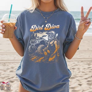 May include: Blue t-shirt with the text "Dirt Diva" in an orange and white font. The graphic features a woman in a helmet and goggles, kneeling near an ATV. The design is in shades of orange, white, and black.