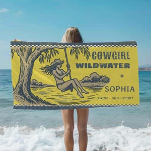 Peut inclure: Serviette de plage jaune avec un dessin de cowgirl sur une balançoire, un arbre et les mots "COWGIRL WILDWATER SOPHIA SWING - SUN - SPIRIT". La serviette a une bordure à carreaux noir et blanc.