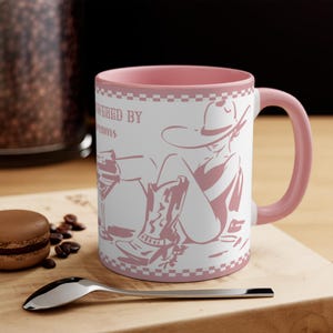 Peut inclure: Mug en céramique blanche avec intérieur et anse roses. L'illustration rose représente une cowgirl avec des bottes et un chapeau, avec l'inscription "POWERED BY DREAMS". Le mug est posé sur une surface en bois.