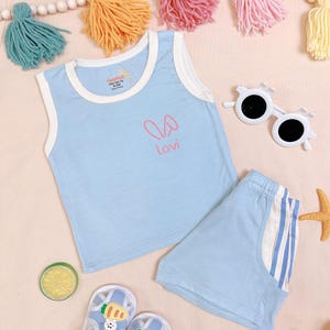 May include: A light blue baby outfit set, including a tank top and shorts. The tank top has a white trim and a pink bunny graphic with the name "Lovi". The shorts have blue and white stripes on the side. The set is for babies 6-12 months.