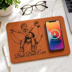 May include: A brown wireless charging pad with a custom engraving of a father and child. The pad has the text "Best Dad Ever" and is charging a smartphone. Tortoiseshell glasses and a pen are also visible.