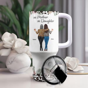May include: White travel mug with a handle and a black lid. The mug features a graphic of two women with the text "Like Mother Like Daughter" and the names "Sophia" and "Olivia". The design includes leopard print and floral accents.