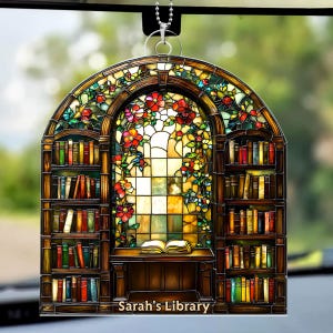 May include: A stained-glass style car hanging ornament featuring a library scene. The design includes bookshelves, a desk with an open book, and a window with floral accents. The text "Sarah's Library" is at the bottom.