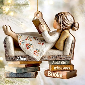 Puede incluir: Adorno de madera de una niña leyendo un libro, reclinada en un sofá. La niña lleva un vestido blanco con un estampado floral. El adorno tiene el texto "Alice" y "Just A Girl Who Loves Books".