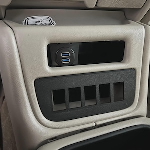 May include: A car interior console with a black panel featuring five rectangular openings. Above, a black module houses two USB ports. The console is beige and has a sticker on top. The overall design suggests an automotive accessory or modification.