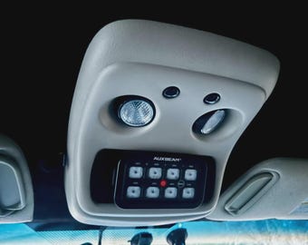 May include: A light-colored vehicle overhead console with integrated lights and a control panel. The control panel, labeled "AUXBEAM", features multiple illuminated buttons. The console is mounted above the windshield, near the sun visors.