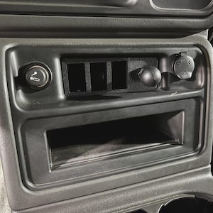 May include: A black car dashboard console featuring a cigarette lighter, three blank switch slots, a 12V power outlet, and a small storage compartment. The console is made of textured plastic.