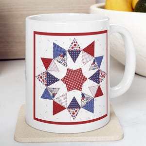 Op de afbeelding: Witte keramische mok met een sterquiltdesign in rood, wit en blauw. Het sterpatroon is omlijst door een rode rand. De mok staat op een beige onderzetter, met een vage achtergrond van een houten oppervlak en een fruitschaal.