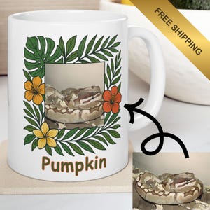 May include: White ceramic mug featuring a framed image of a snake, surrounded by green leaves and orange and yellow flowers. The word "Pumpkin" is printed in brown. A small image of the snake is in the lower right corner.