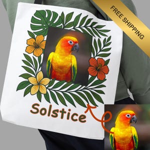 Peut inclure: Sac fourre-tout blanc avec un perroquet jaune, orange et vert encadré de feuilles et de fleurs vertes. Le mot "Solstice" est imprimé en marron. Une bannière dorée avec "LIVRAISON GRATUITE" est visible.